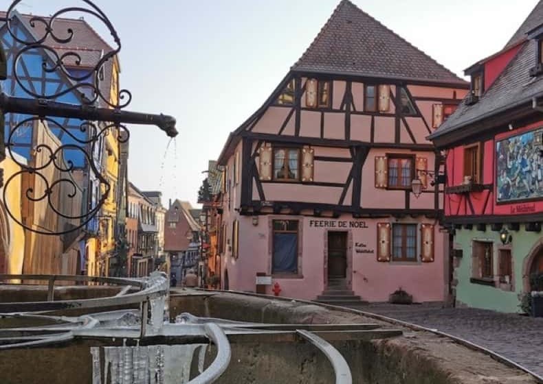 A stone fountain in a village square below painted house fronts and a wrought-iron shop sign