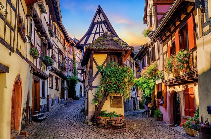 A cobbled village lane at sunset between half-timbered fronts, an old wine press under the gable in the middle of it